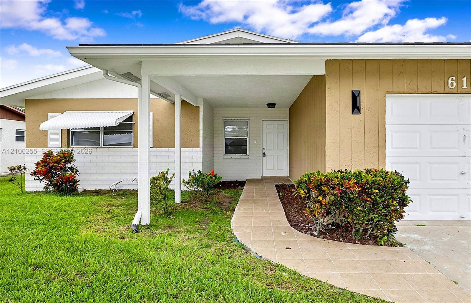 6118 Dusenburg Road, Delray Beach, FL 33484 Exterior facade featuring a combination of stucco and painted brick, covered entryway with a tiled walkway, white sectional garage door, and metal window awnings