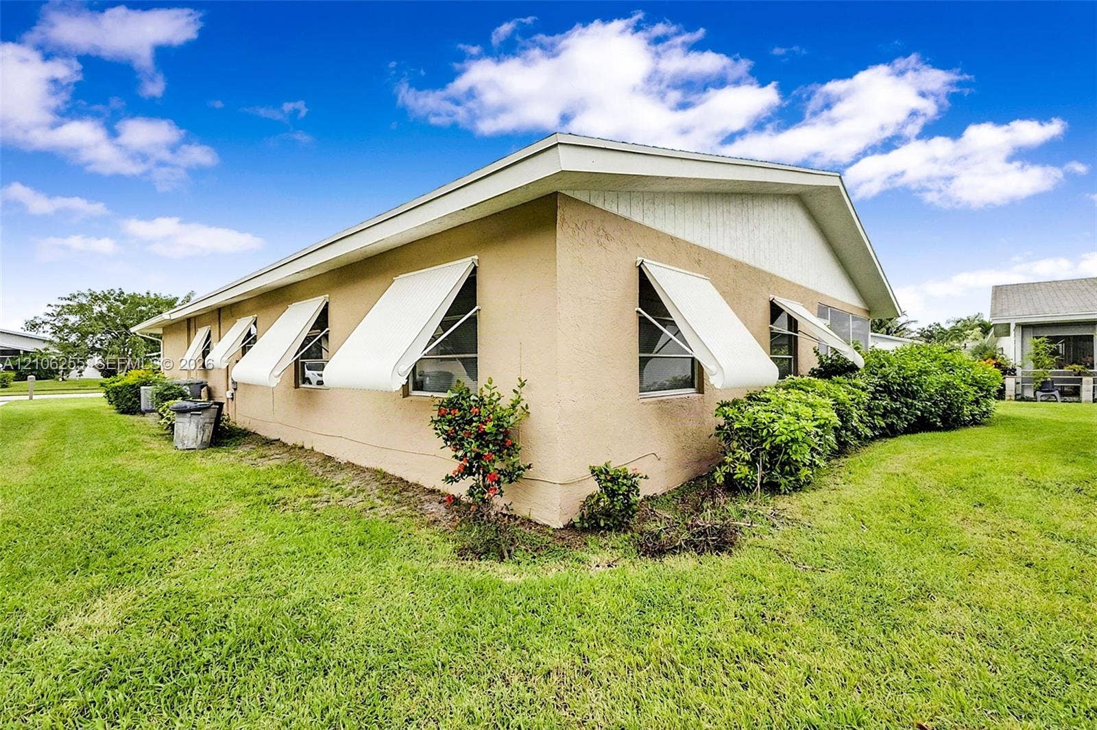 6118 Dusenburg Road, Delray Beach, FL 33484 Exterior of a stucco-finish residence featuring a gabled roofline, retractable white window awnings, and low-maintenance landscaping set within a sprawling grass lawn