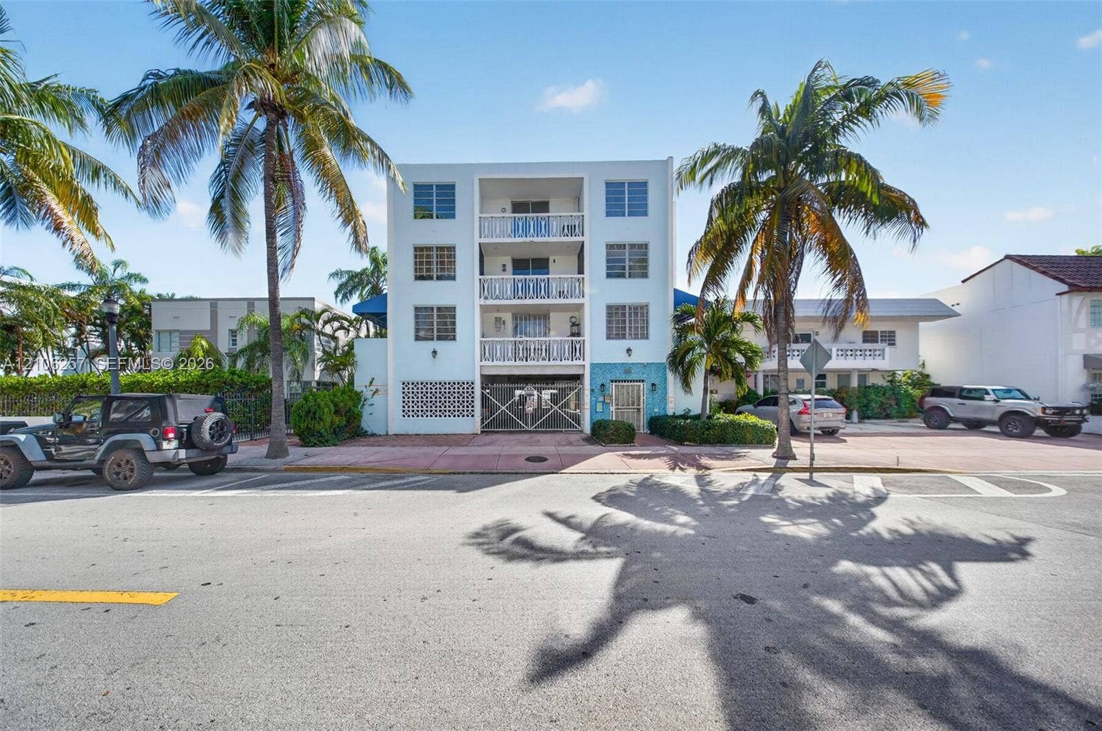 Multi-story residential building exterior featuring stacked balconies with patterned railings, decorative textured tile facade, louvered windows, and mature palm trees