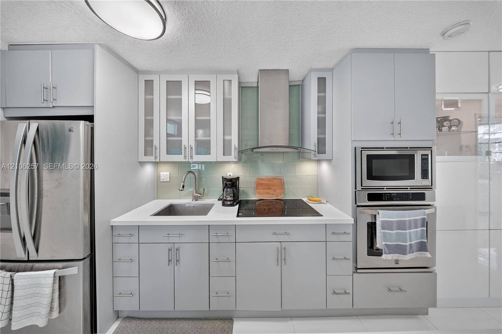 Kitchen featuring light gray cabinetry with glass-front uppers, solid-surface countertops, a glass tile backsplash, and stainless steel built-in appliances including a wall-mounted range hood