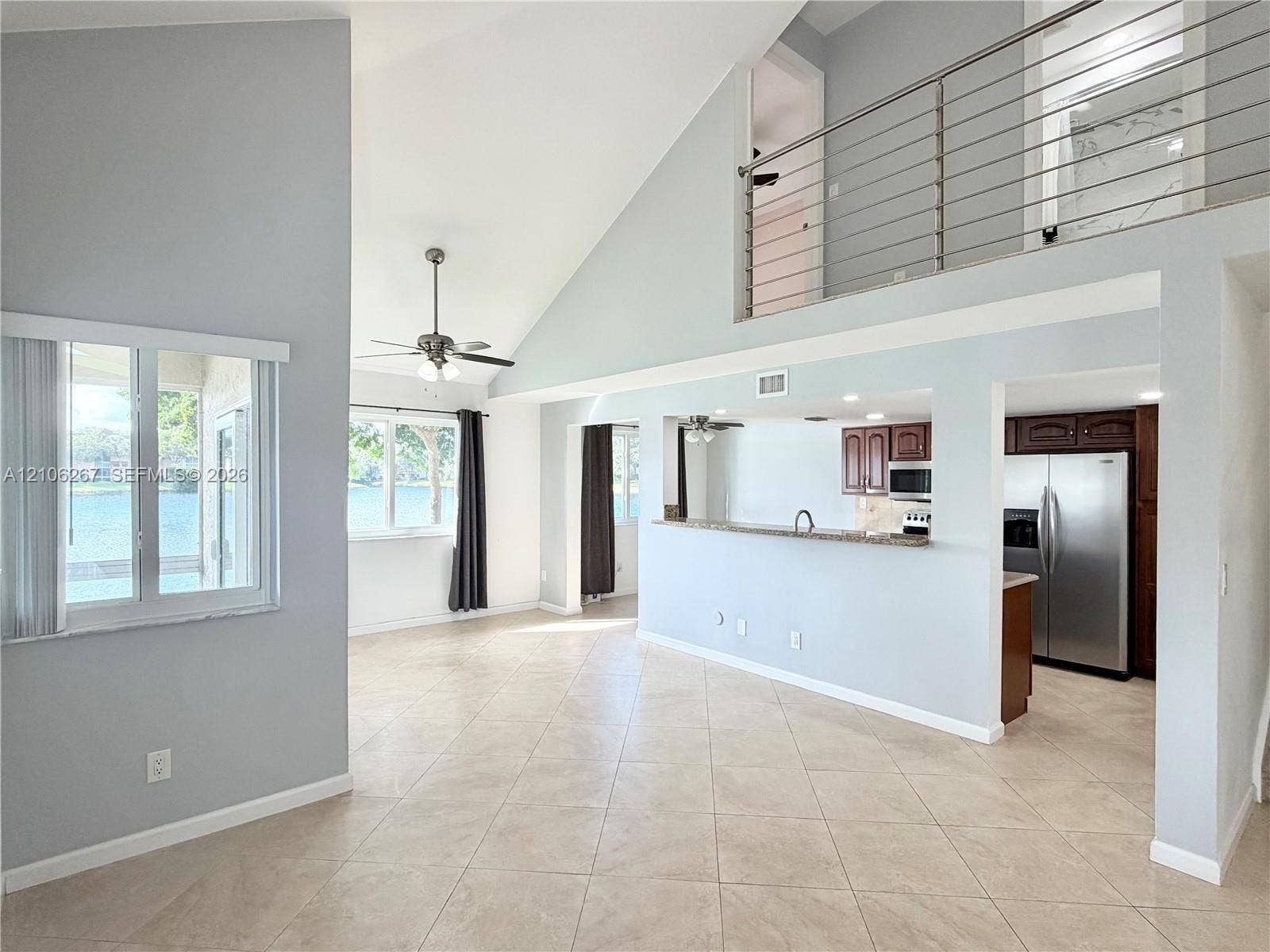 Vaulted ceilings, tiled flooring, metal horizontal-bar staircase railing, kitchen pass-through window with bar seating, and large windows overlooking water
