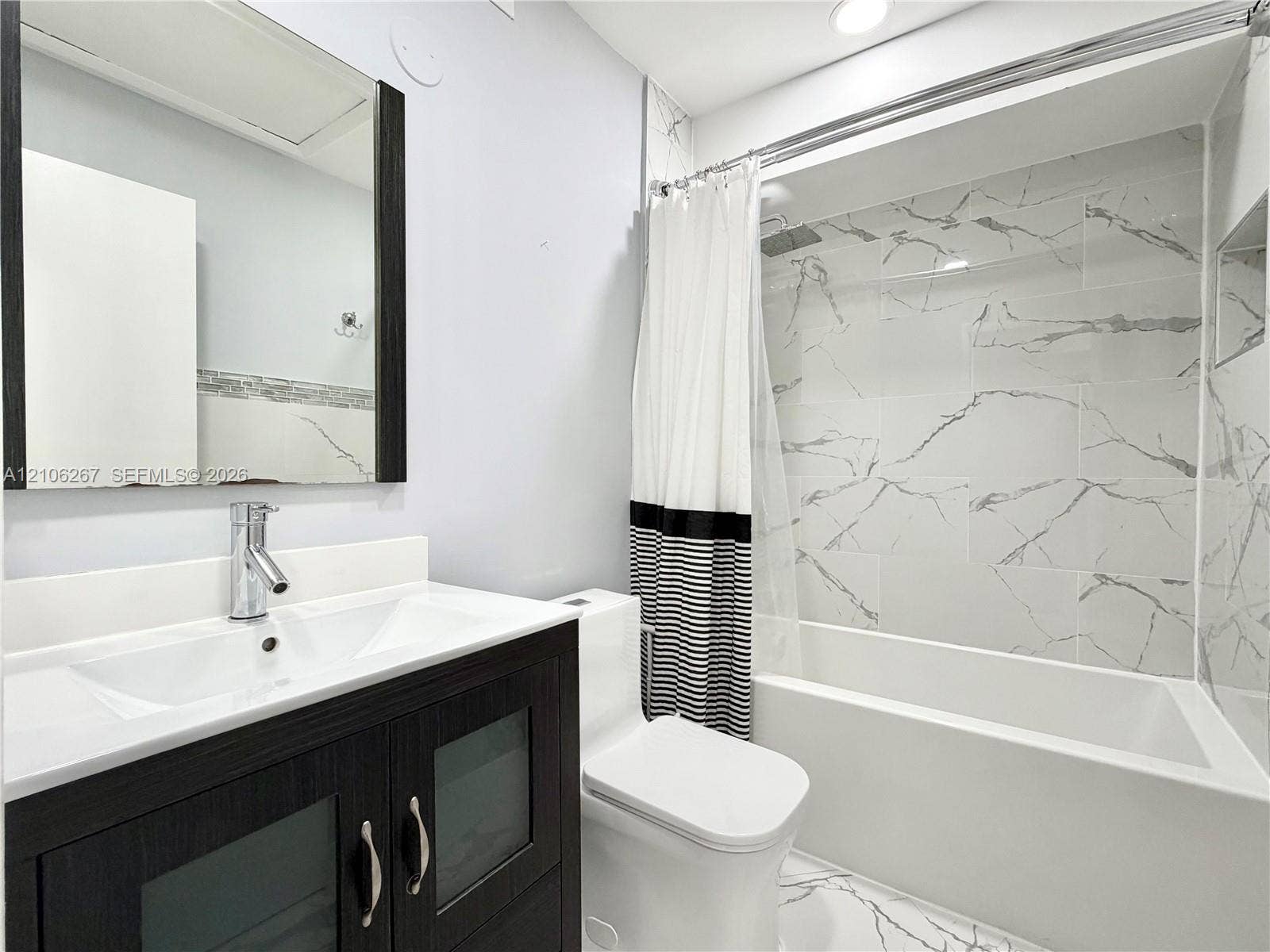 Bathroom featuring a dark wood-finish vanity with frosted glass inserts, a white integrated countertop sink, a full-length framed mirror, and a tub-shower combination with veined tile surround