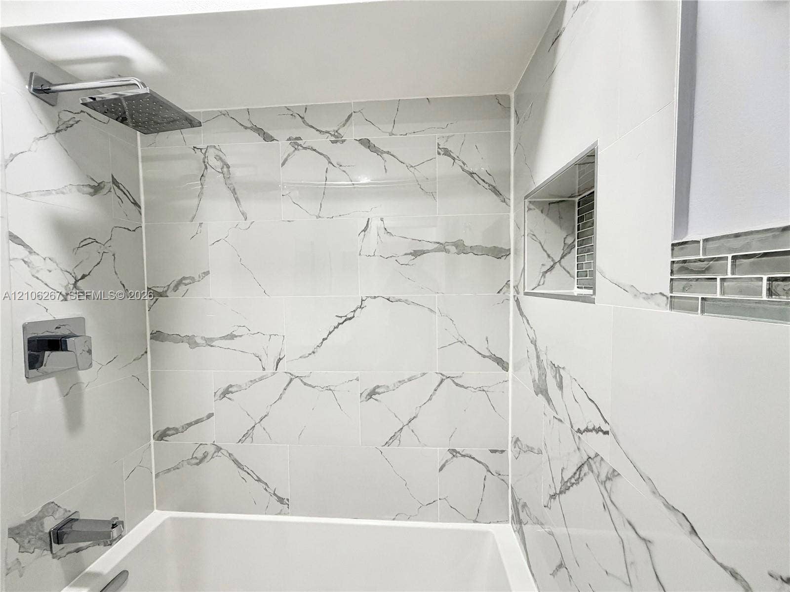 Shower area featuring large-format veined tile surround, recessed shelving niche, glass tile accent border, and chrome rainfall showerhead