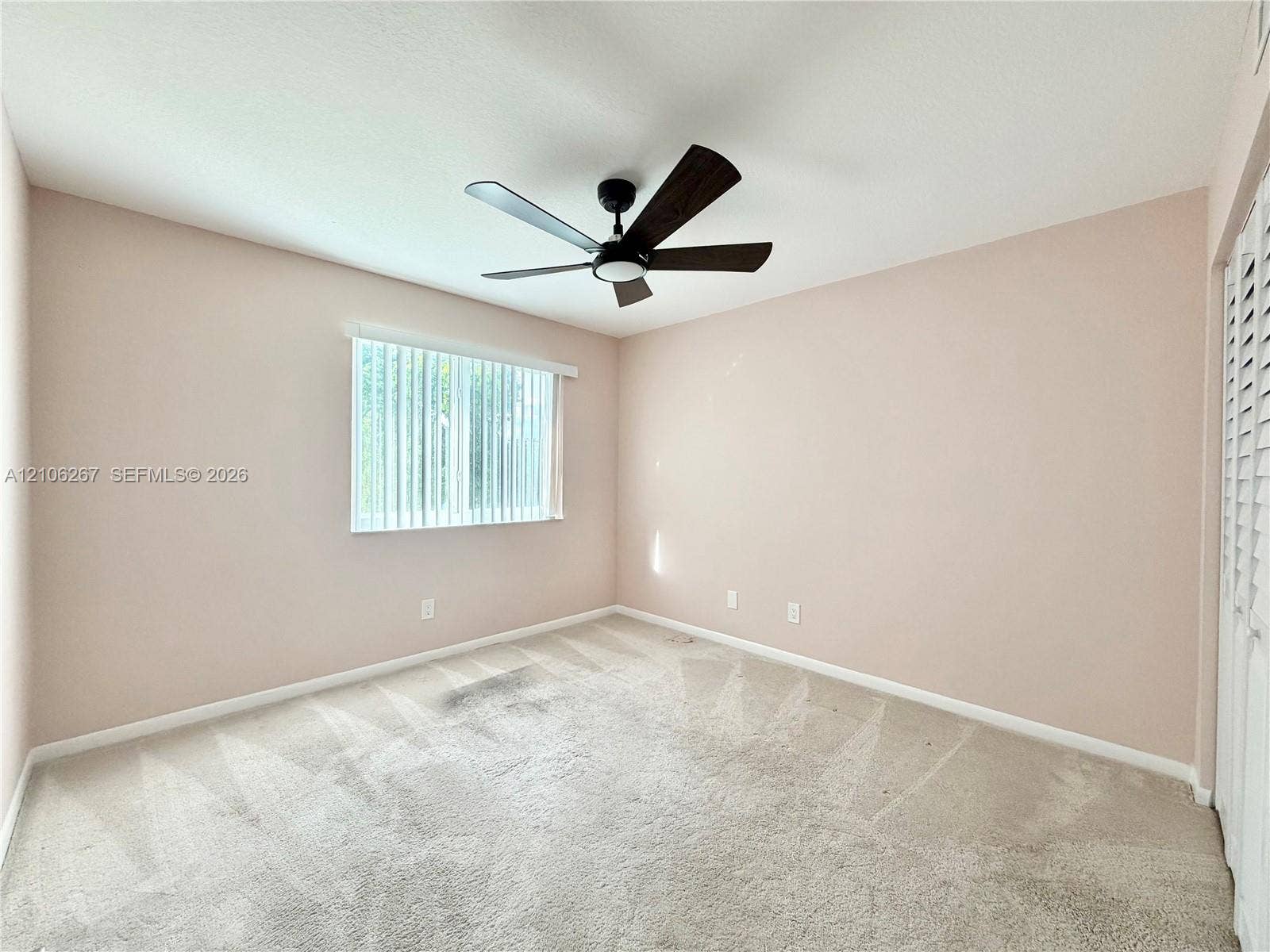 Interior space featuring neutral wall tones, wall-to-wall carpeting, a ceiling fan with integrated lighting, a window with vertical blinds, and bifold closet doors