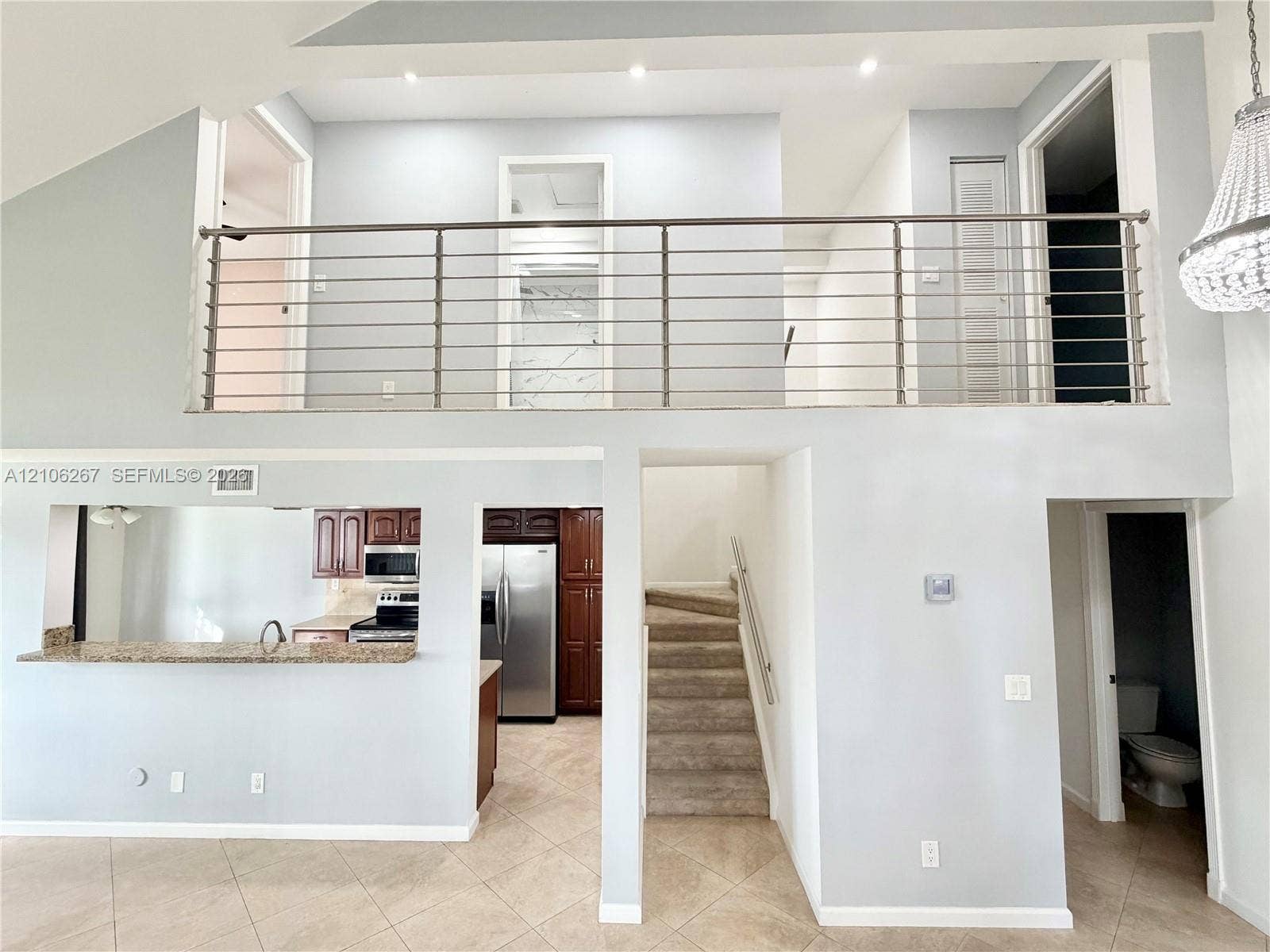 Vaulted ceiling, metal horizontal bar railing, neutral tile flooring, kitchen pass-through with stone countertop, and crystal-style chandelier