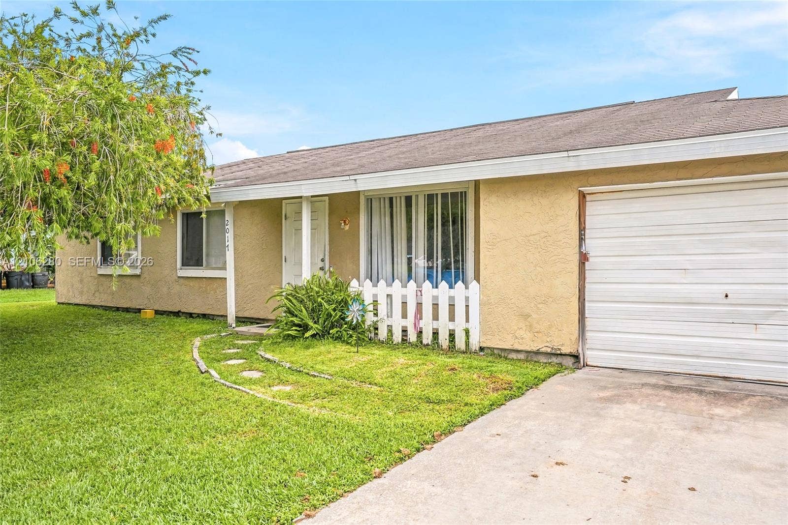 2017 SE Ancora Court, Port St Lucie, FL 34952 Single-story stucco exterior featuring a concrete driveway, attached garage, stone-paver walkway, and white picket fencing