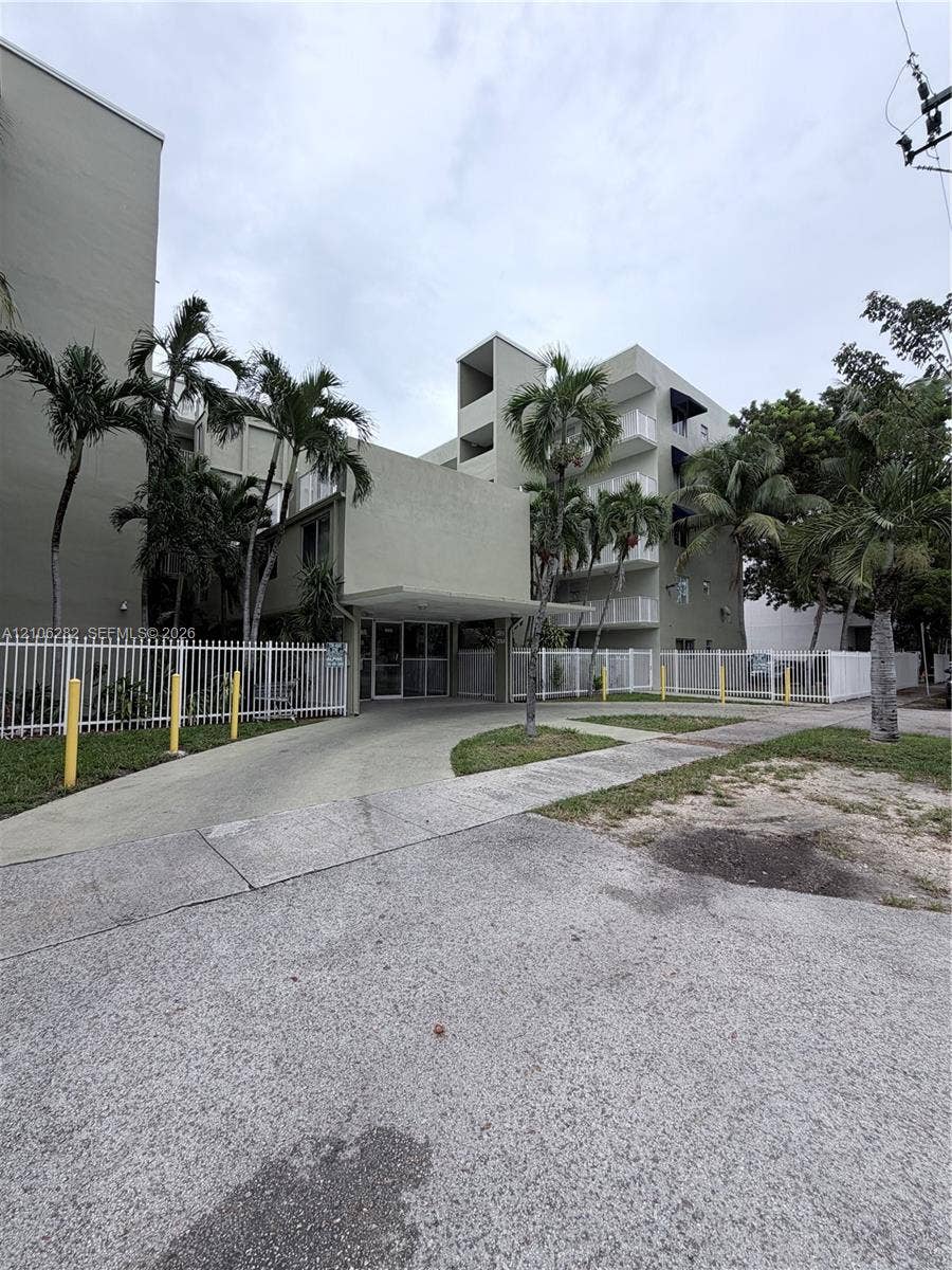 665 NE 83rd Terrace, #210, Miami, Miami, FL 33138 Multi-story building exterior featuring a covered entryway, concrete driveway, white metal fencing, and mature palm trees