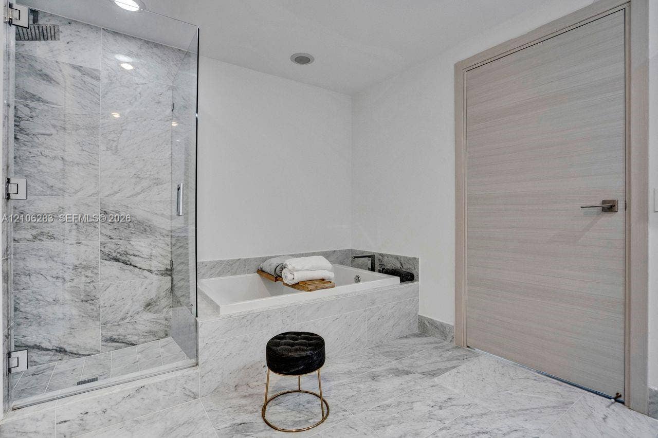 801 S Miami Avenue, #1801, Miami, FL 33130 Frameless glass shower enclosure with stone tile surround, built-in soaking tub, stone tile flooring, and a light-toned wood-finish interior door