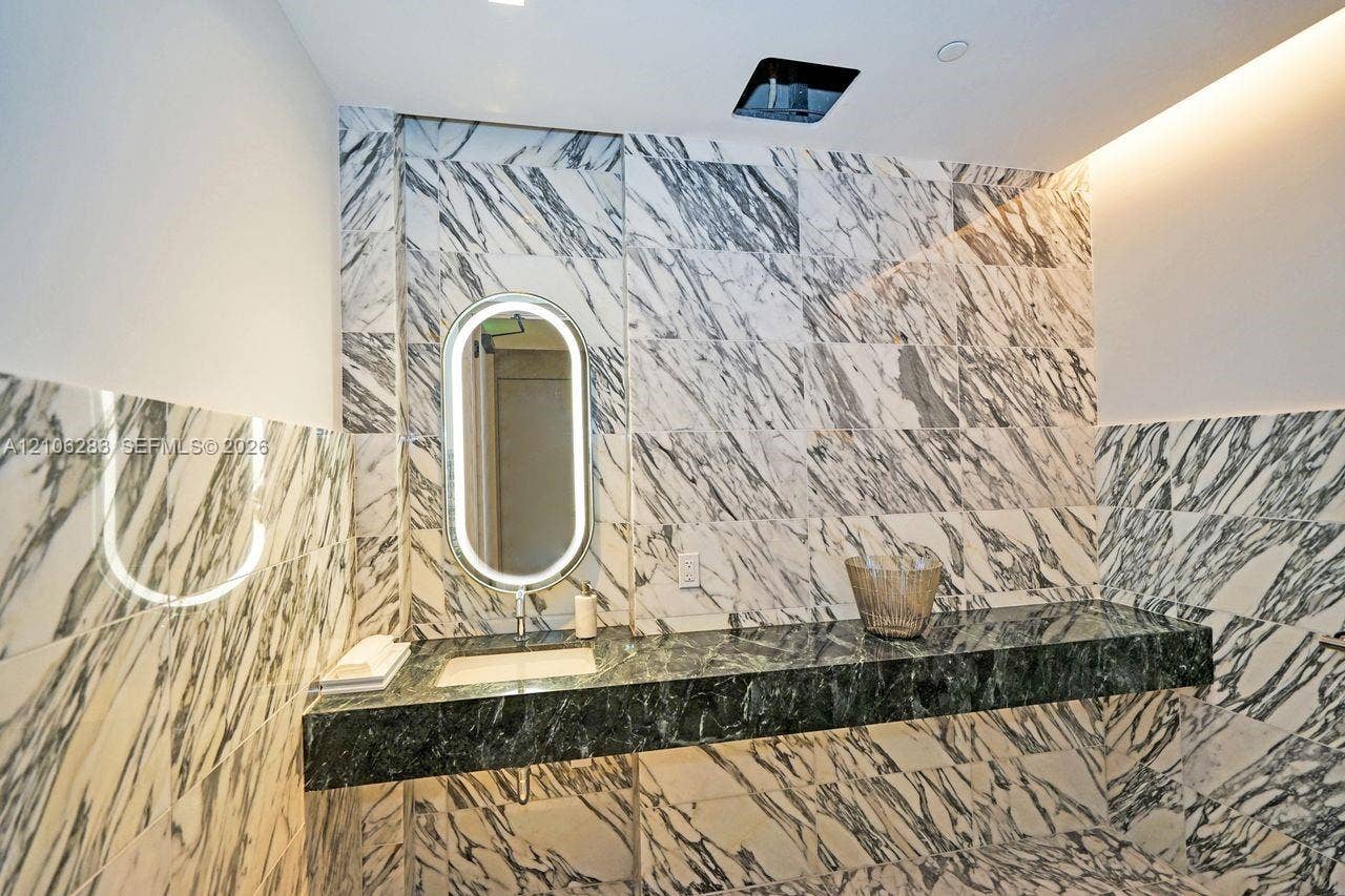 801 S Miami Avenue, #1801, Miami, FL 33130 Floating vanity with a dark natural stone countertop, integrated sink, illuminated oval mirror, and floor-to-ceiling veined stone tile wall covering