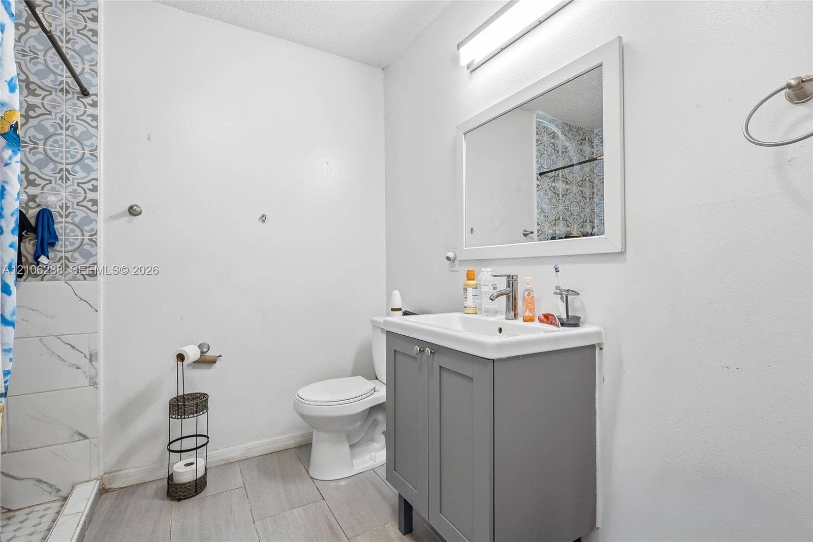 Bathroom featuring a grey shaker-style vanity with an integrated basin, large rectangular vanity mirror, patterned tile wall accents in the shower, and neutral floor tile