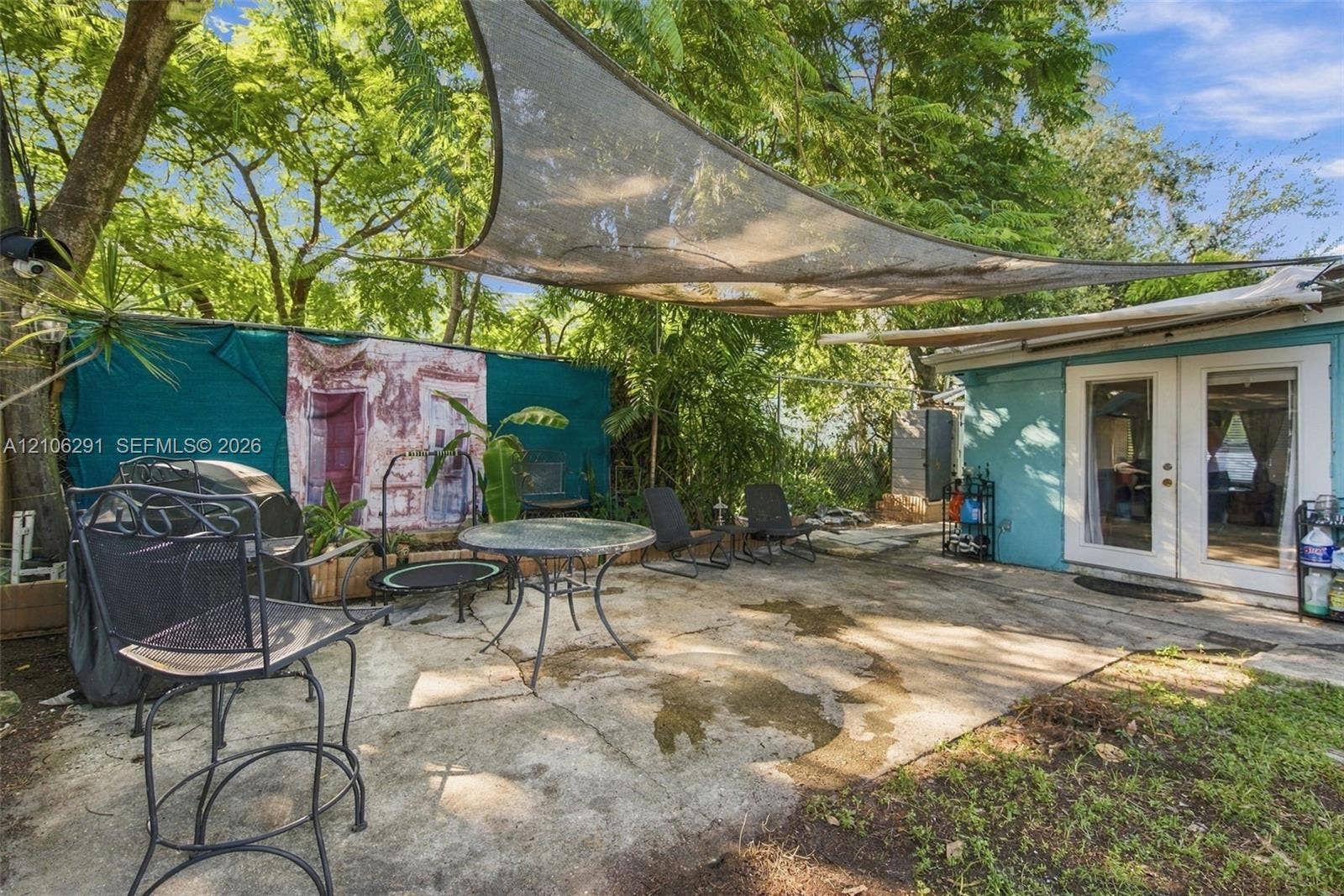 Concrete patio featuring a retractable shade sail, mature trees, and dual glass-paned doors