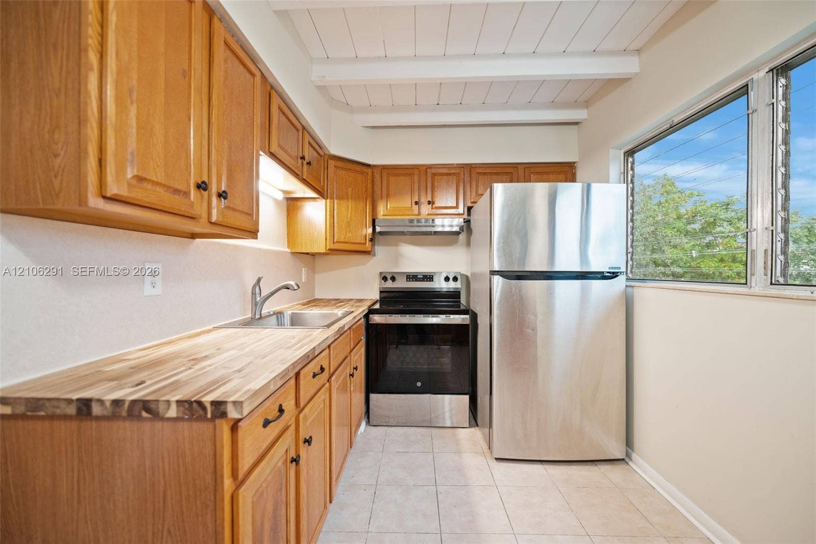 Kitchen featuring stained wood cabinetry, butcher-block countertops, stainless steel appliances, tile flooring, and a ceiling with exposed white beams