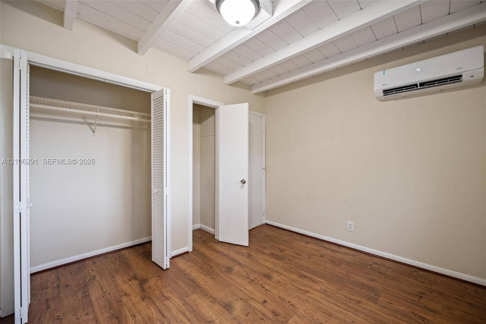 Room featuring wood-finish flooring, exposed painted ceiling beams, a wall-mounted ductless air conditioning unit, and dual closet storage options