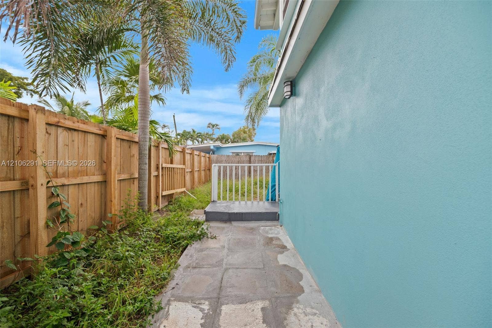 Exterior side yard featuring a smooth stucco finish, stone paver walkway, wood privacy fencing, and exterior wall-mounted lighting