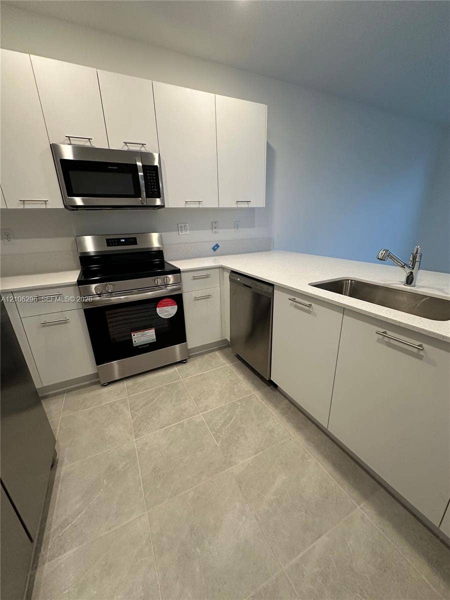 12103 NW 24th Place, #12103, Miami, Miami, FL 33167 Kitchen featuring flat-panel white cabinetry, light-toned stone-finish countertops, stainless steel appliances, a stainless steel undermount sink with a gooseneck faucet, and large-format tile flooring