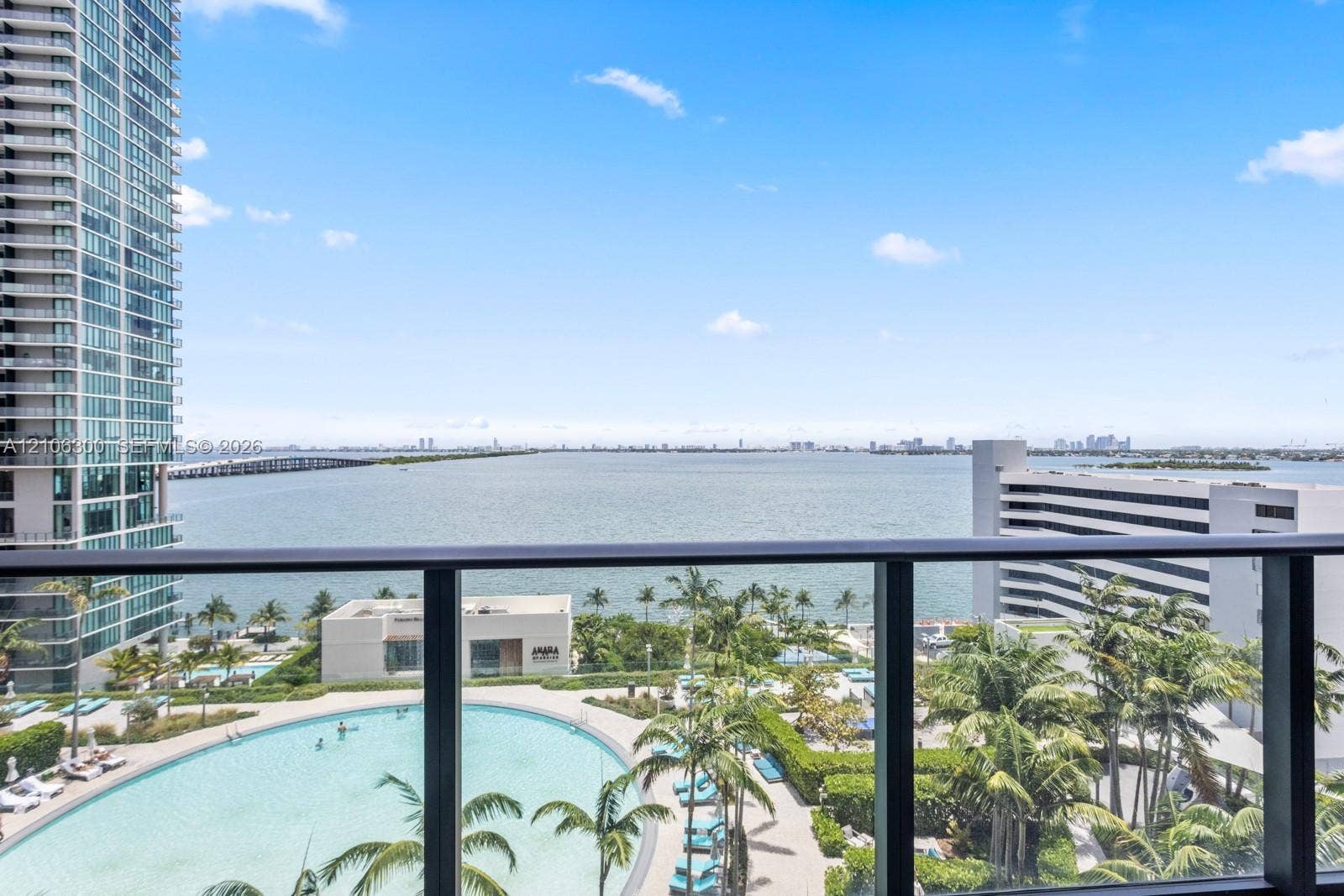 480 NE 31st Street, #1005, Miami, Miami, FL 33137 Balcony featuring a dark metal railing, overlooking a large swimming pool, landscaped grounds with mature palm trees, and coastal water