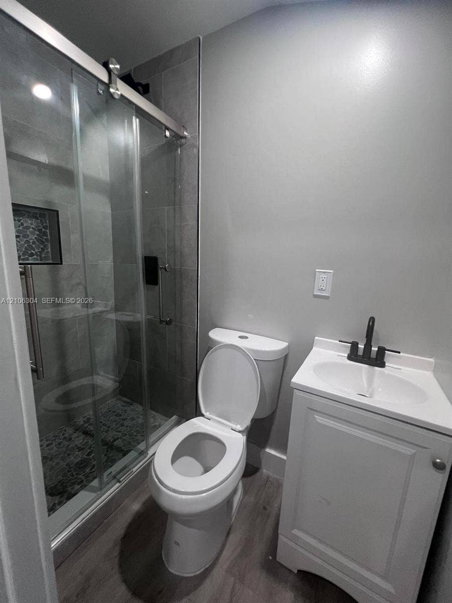 Glass-enclosed shower with tiled surround, wood-finish flooring, vanity with integrated basin, and matte black plumbing fixtures
