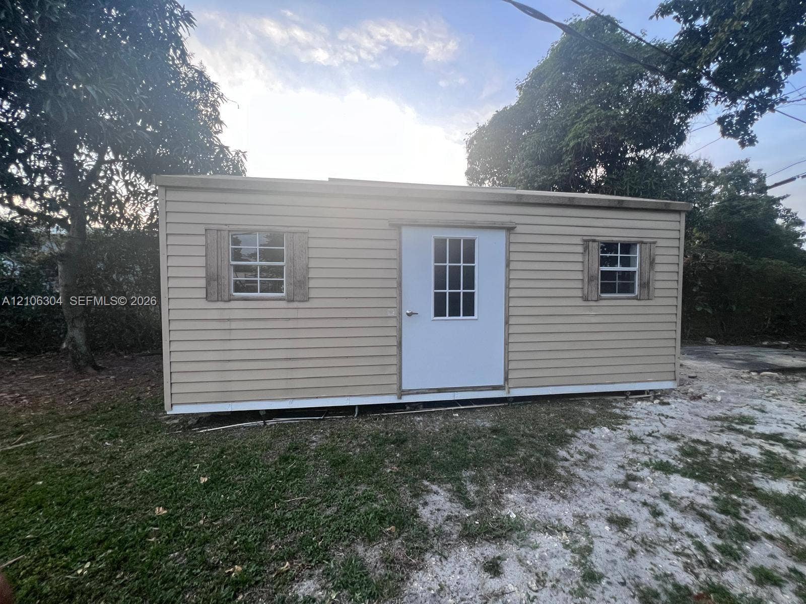 Outbuilding featuring horizontal lap siding, a central entry door with window pane detailing, two exterior windows with wood-tone shutters, and a flat roofline