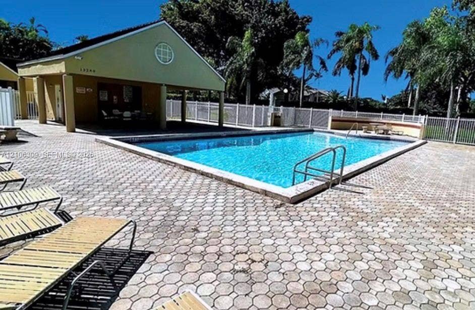 8888 SW 131st Court, #409, Miami, FL 33186 In-ground swimming pool with metal ladder entry, surrounded by a paved deck featuring stone-style pavers, enclosed by a perimeter fence, and accompanied by a covered pavilion structure