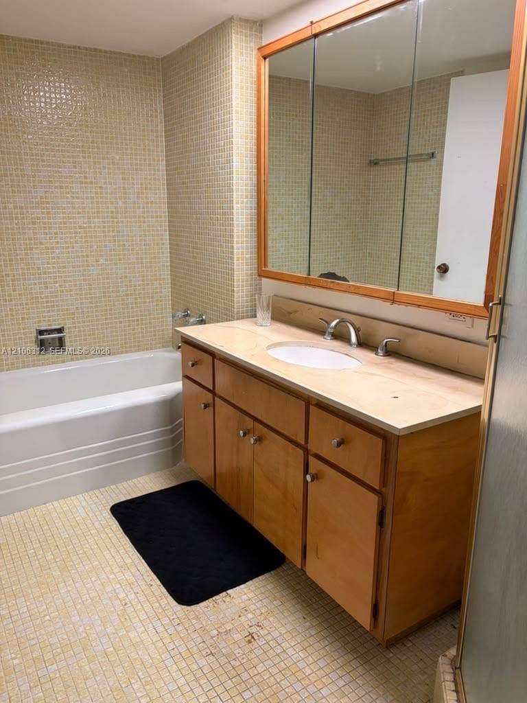 800 NE 195th Street, #311, Miami, Miami, FL 33179 Bathroom featuring a wood-finish vanity with a neutral countertop, undermount oval sink, three-panel medicine cabinet with wood trim, and wall-to-wall mosaic tile detailing around the built-in bathtub