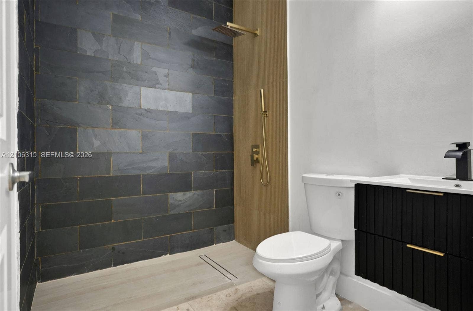 2319 NE 35th Drive, Fort Lauderdale, FL 33308 Walk-in shower featuring dark stone-style tile, gold-finish hardware, and a wood-finish panel accent, paired with a black floating vanity and integrated sink