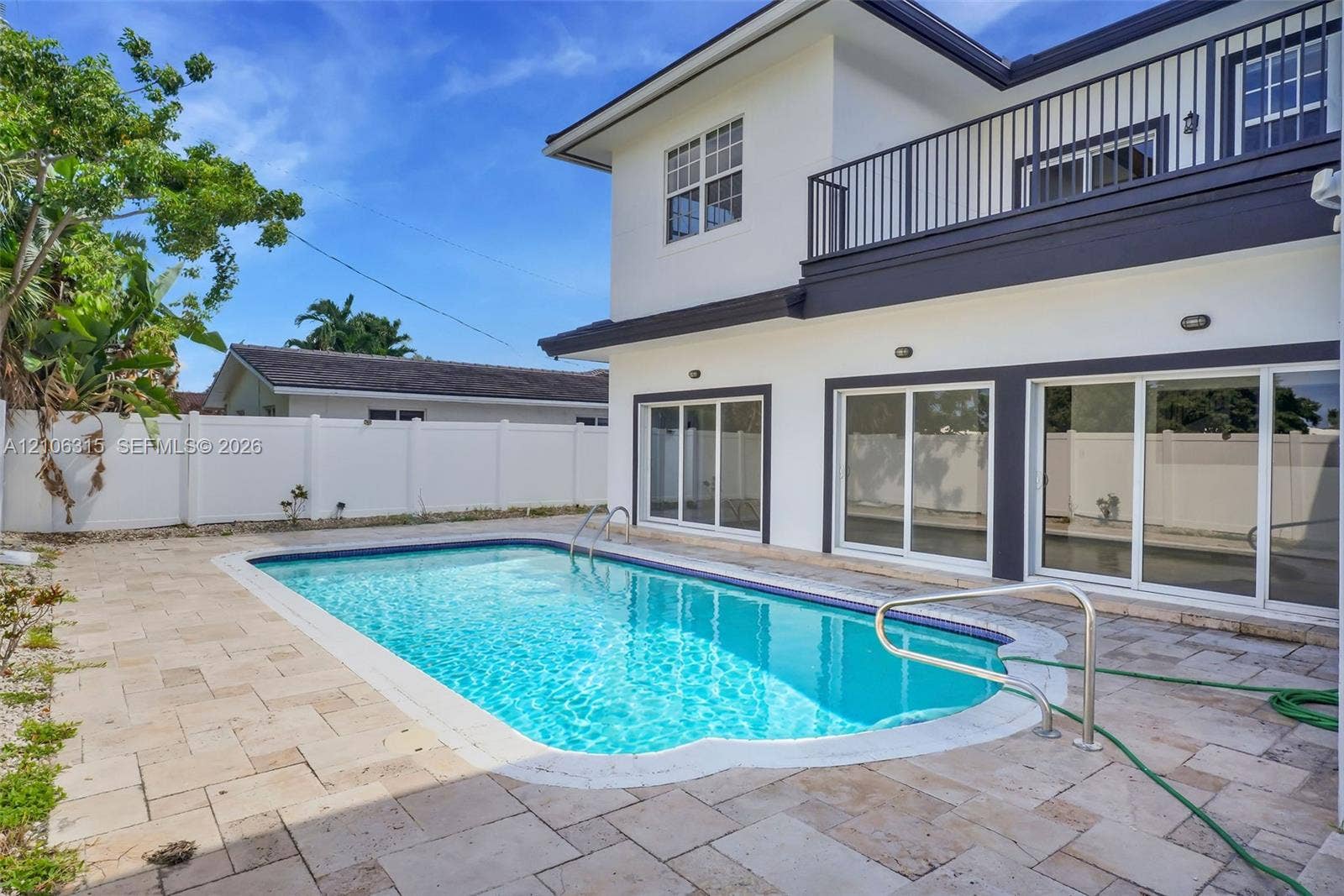 2319 NE 35th Drive, Fort Lauderdale, FL 33308 Exterior featuring a white stucco facade, private swimming pool, stone paver decking, white vinyl perimeter fencing, and an upper-level balcony with metal railing