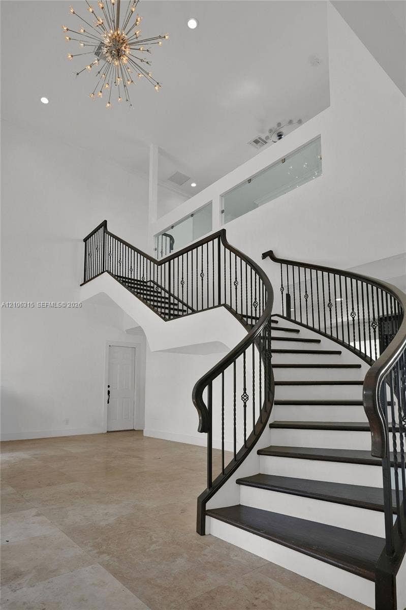 2319 NE 35th Drive, Fort Lauderdale, FL 33308 Curved staircase featuring dark-finished treads, white risers, and decorative metal balusters, complemented by high ceilings, a starburst chandelier, and neutral stone-finish flooring