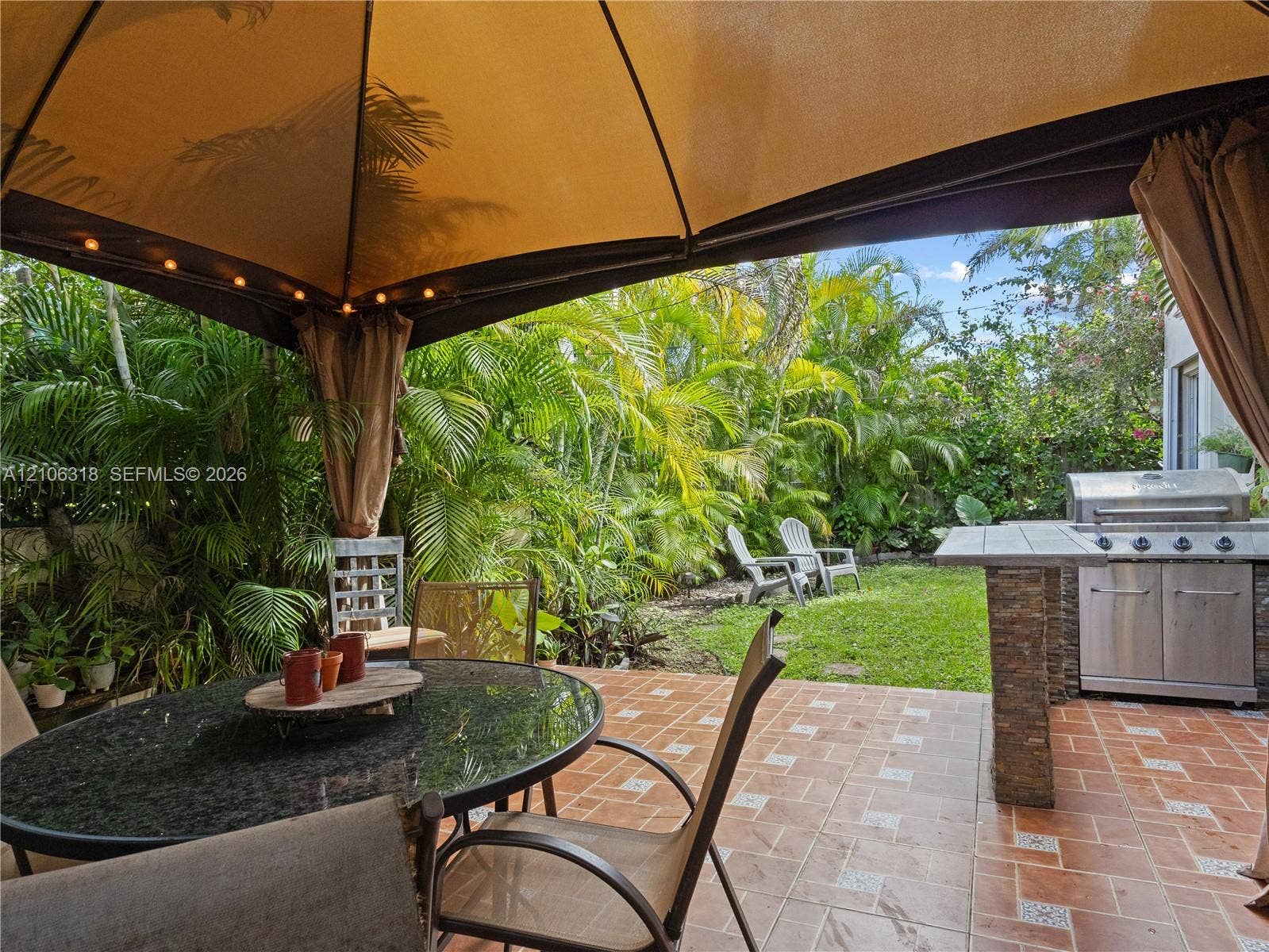 9921 NW 51st Lane, Doral, FL 33178 Outdoor patio featuring terracotta-style tile flooring, built-in outdoor kitchen with stainless steel grill, stone-finish base, and landscaped backyard with mature greenery
