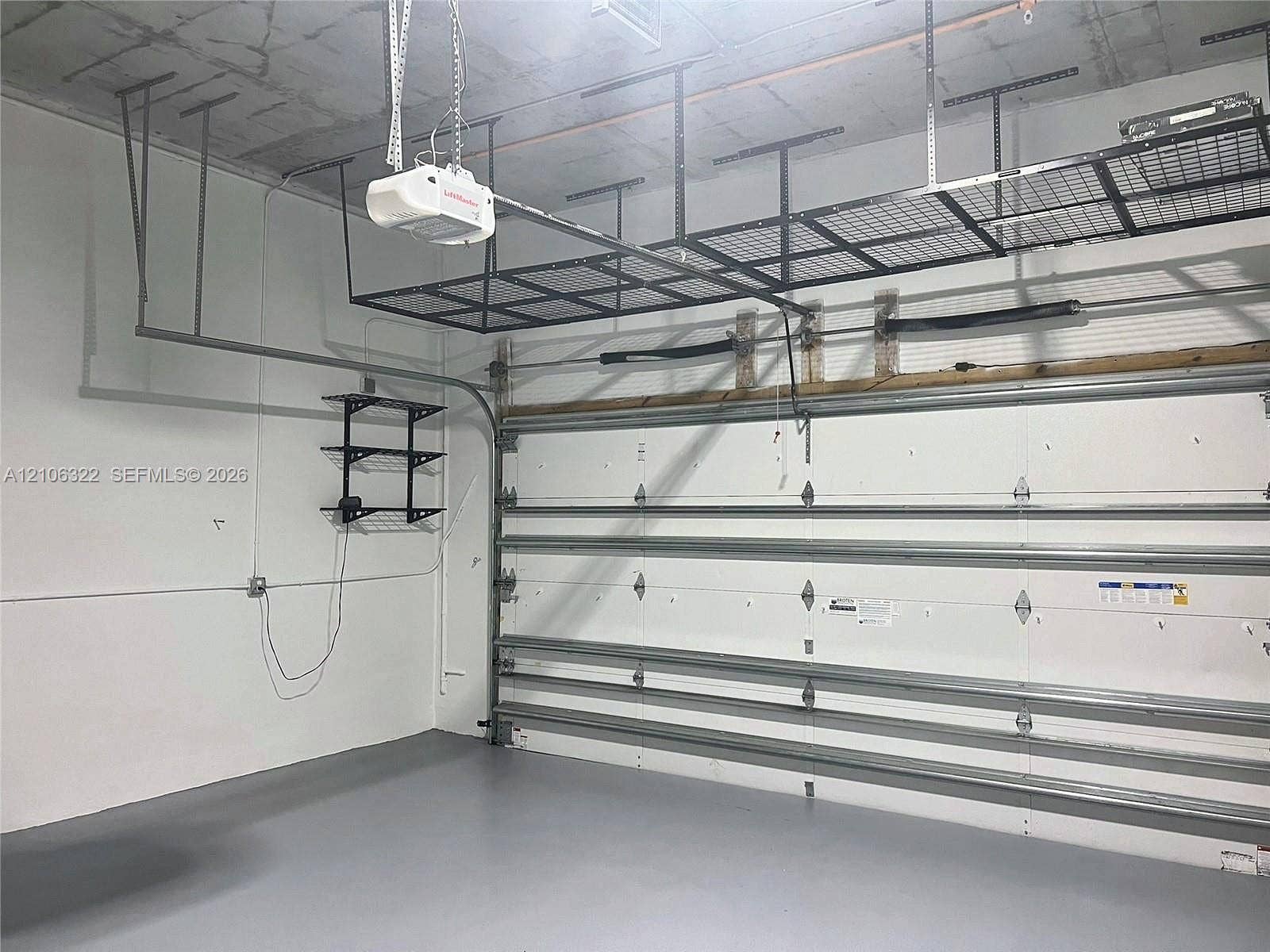 Garage space featuring finished epoxy flooring, white paneled walls, exposed concrete ceiling, integrated overhead metal storage racks, and wall-mounted metal shelving