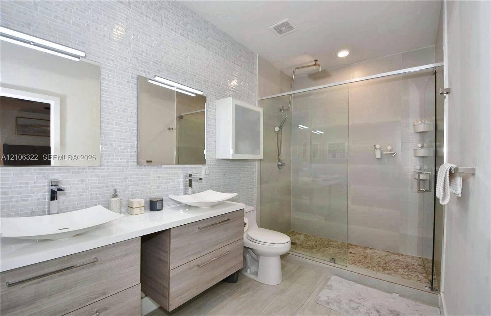 Dual floating vanity with rectangular vessel sinks, chrome faucets, wall-mounted medicine cabinet, and a walk-in glass-enclosed shower featuring a textured stone-finish floor