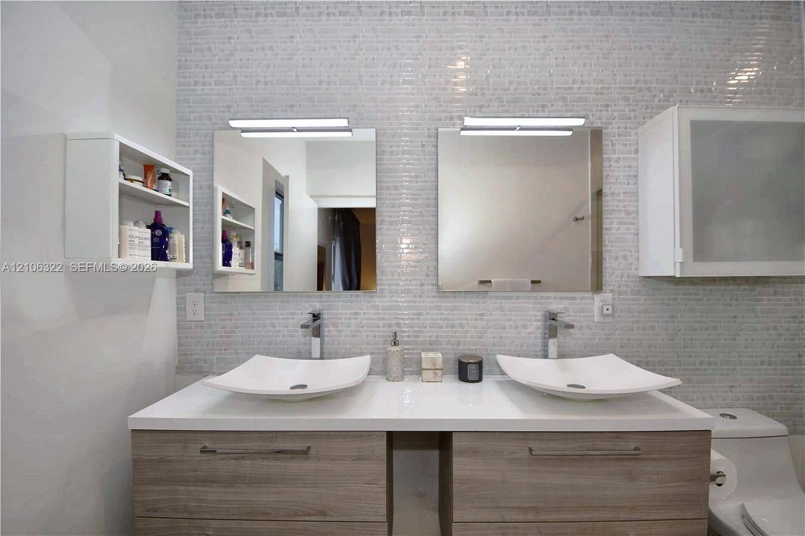 Double vanity with wood-finish cabinetry, white countertop, dual vessel sinks, chrome faucets, and tiled wall backsplash