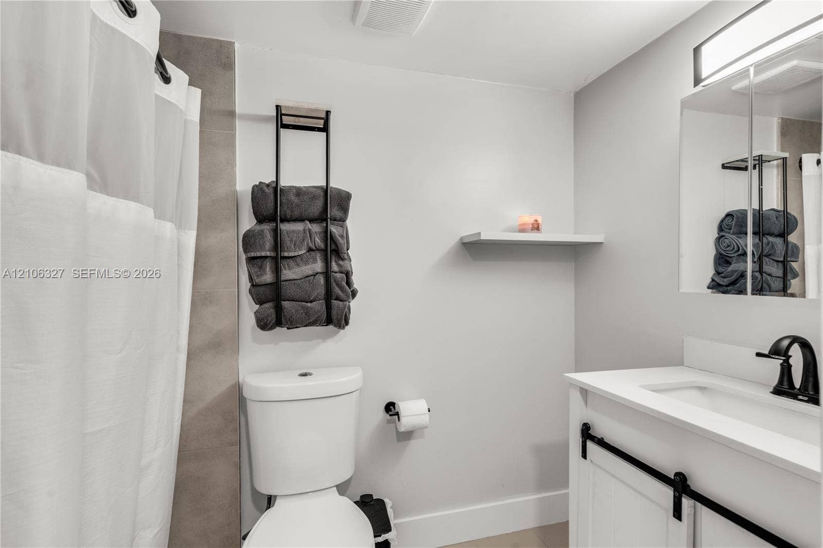 Bathroom featuring a white vanity with a composite countertop, matte black faucet, mirrored medicine cabinet, wall-mounted shelving, and a tiled shower surround