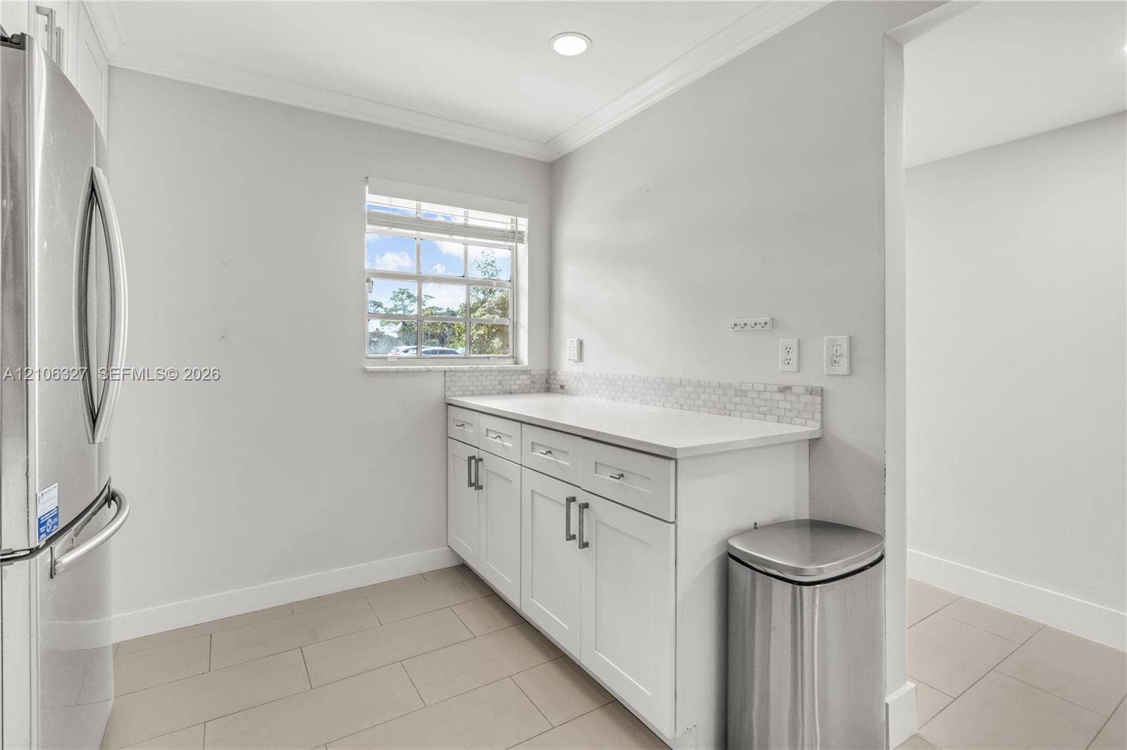 Shaker-style white cabinetry featuring chrome hardware, solid-surface countertop, mosaic tile backsplash, stainless steel refrigerator, and ceramic tile flooring