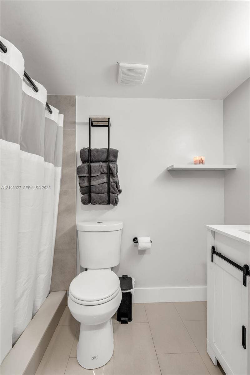 Bathroom featuring large-format neutral tile flooring, white vanity with matte black hardware, integrated tub surround with neutral accent tiling, and a wall-mounted floating shelf