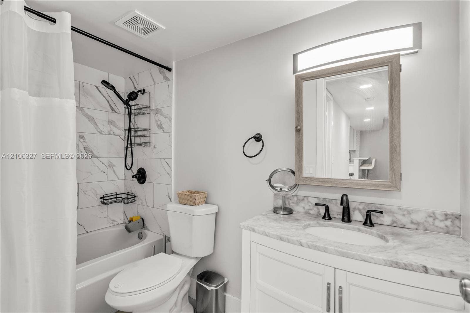 Bathroom featuring a stone-finish countertop with an integrated oval basin, white shaker-style cabinetry, a light-toned wood-frame medicine cabinet, and a bathtub enclosure with marble-patterned wall tile and a matte black shower fixture
