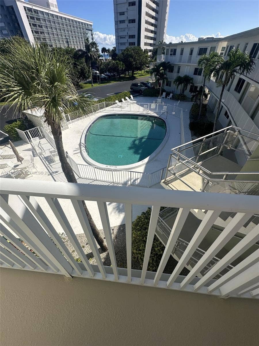 3600 N Ocean Drive, #423, Riviera Beach, Riviera Beach, FL 33404 Elevated balcony with a white metal railing overlooking a community pool area with a paved deck, perimeter fencing, and mature palm trees