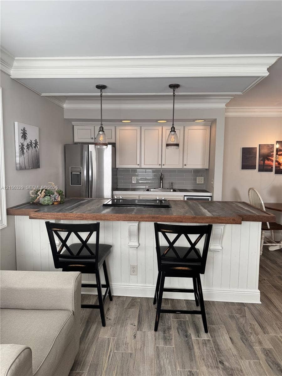 3600 N Ocean Drive, #423, Riviera Beach, Riviera Beach, FL 33404 Kitchen featuring a wood-topped peninsula with integrated cooktop, white cabinetry, subway tile backsplash, stainless steel refrigerator, and dual pendant lighting under crown-molded tray ceiling details