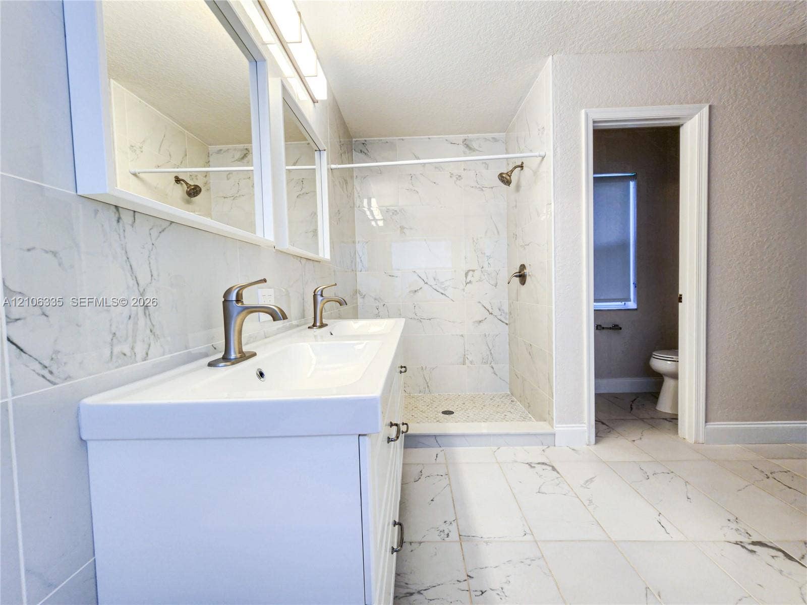 Double vanity with integrated sinks, metallic finish faucets, large wall-mounted mirror, walk-in shower with stone-patterned tile surround, and stone-patterned tile flooring