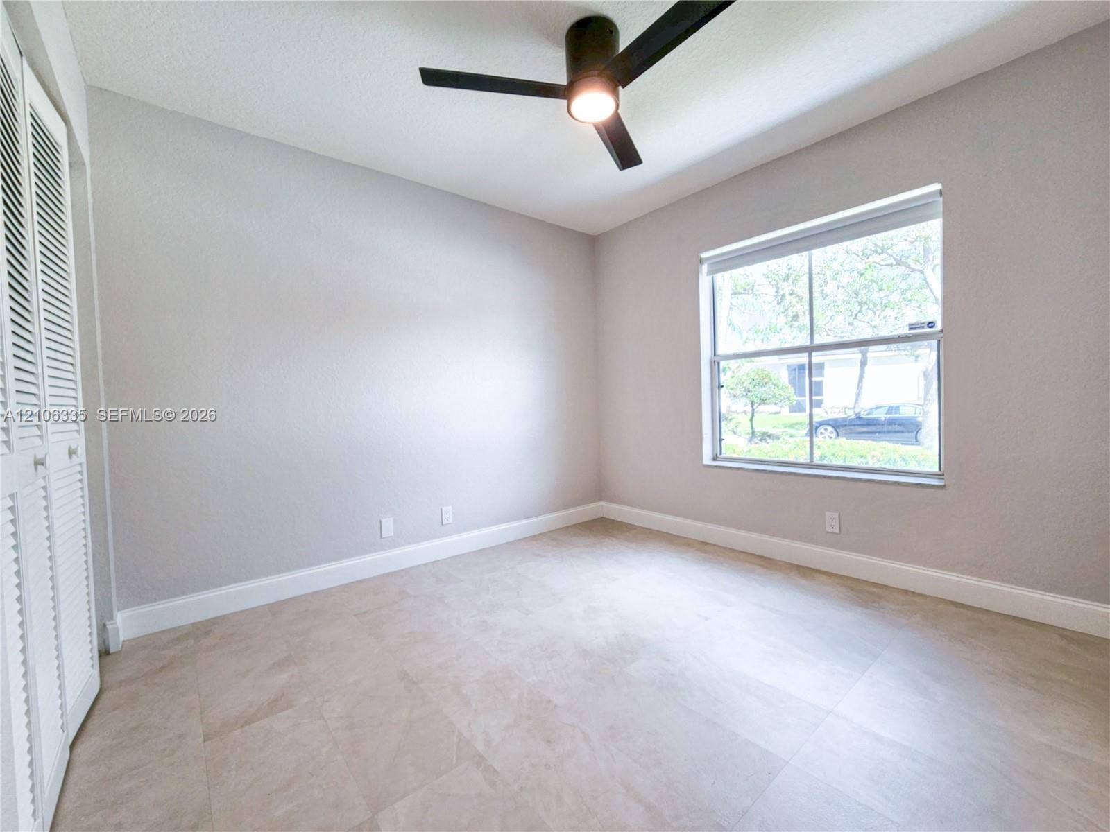 Neutral-toned room featuring large-format tile flooring, a matte black ceiling fan with an integrated light, louvered closet doors, and a single-pane window