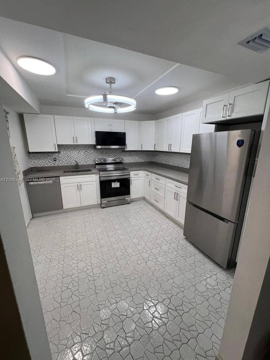 Kitchen featuring white shaker-style cabinetry, stainless steel appliances, patterned tile backsplash, solid-surface countertops, and geometric tile flooring