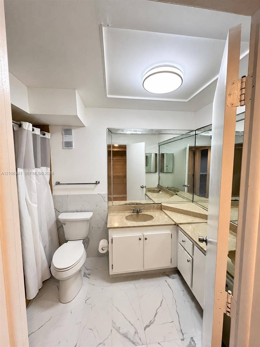 Bathroom featuring white cabinetry, stone-surface countertop, full-width wall mirror, and large-format tile flooring with veining detail