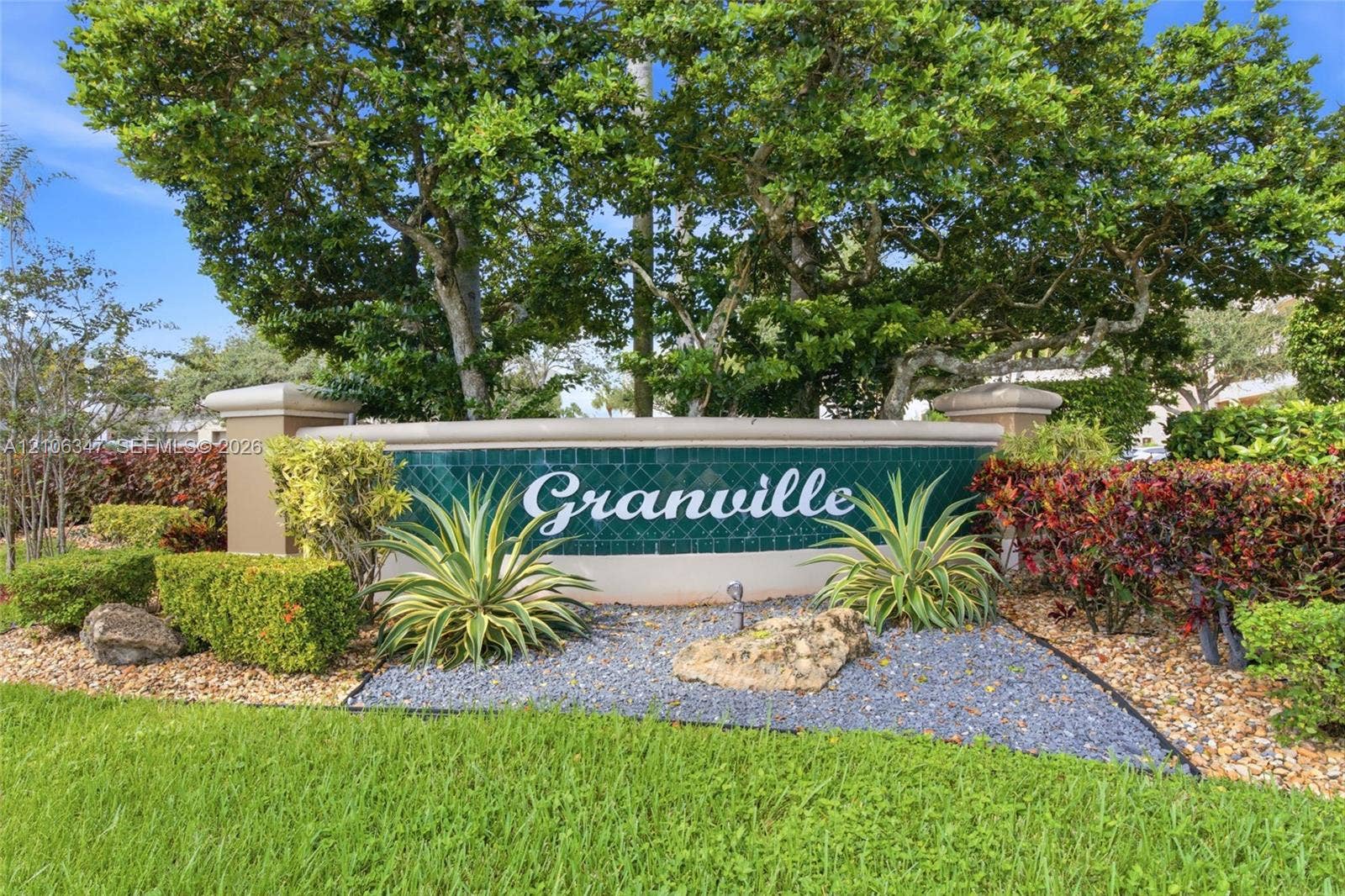 7759 Granville Drive, #306, Tamarac, FL 33321 Community signage featuring emerald green tile accents, stone-finished pillars, and manicured landscaping with mature trees