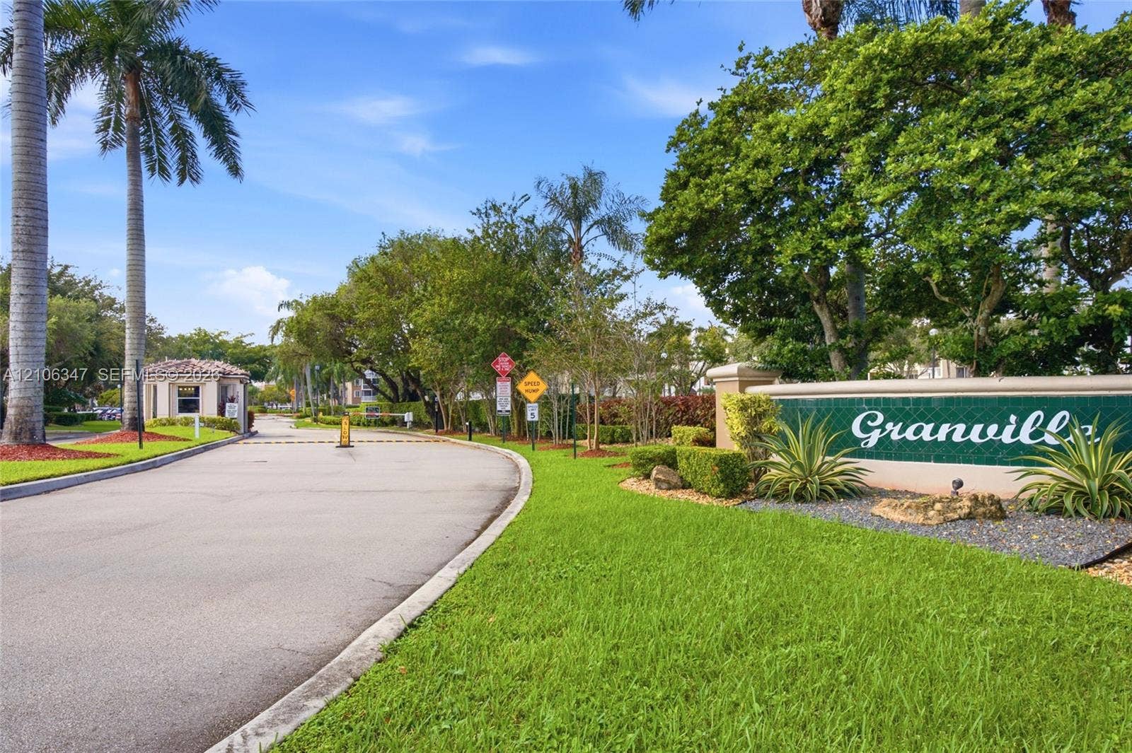7759 Granville Drive, #306, Tamarac, FL 33321 Gated community entrance featuring paved access, a security booth, manicured lawns, mature palm trees, and professional landscaping