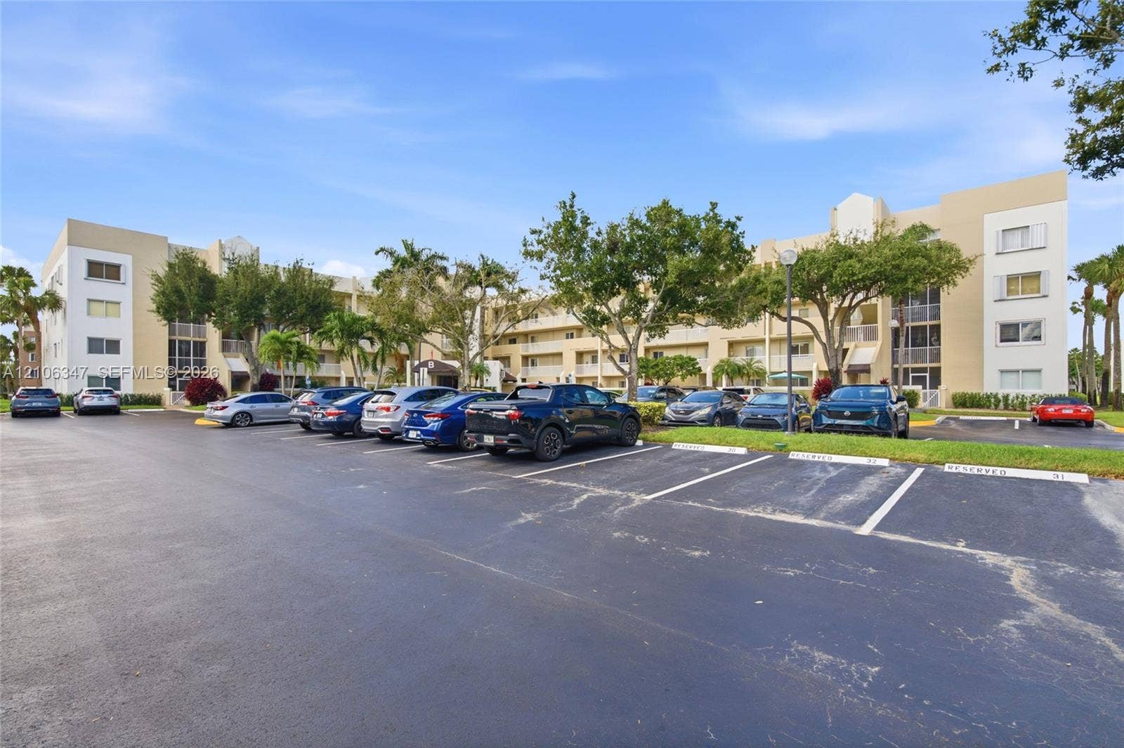 7759 Granville Drive, #306, Tamarac, FL 33321 Multi-story residential building exterior featuring a neutral stucco facade, inset balconies, paved surface parking lot, and mature palm trees