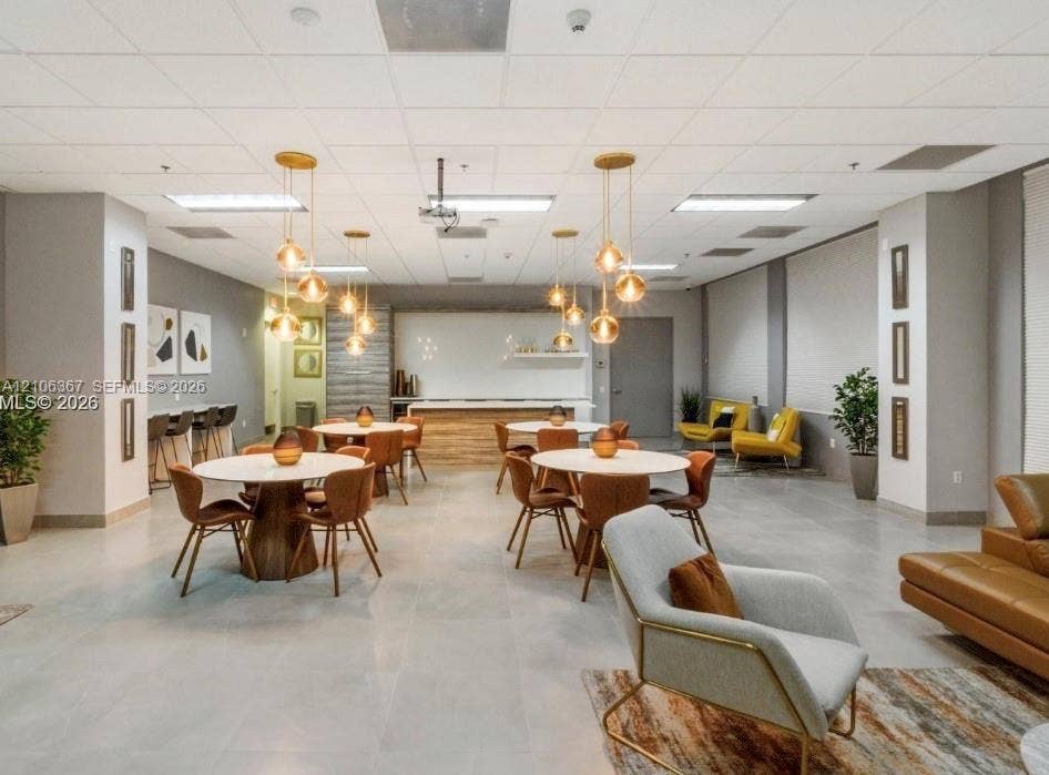 Open-concept common area featuring large-format tile flooring, a built-in refreshment bar with quartz-style countertops, drop ceiling grid, and multiple gold-tone pendant light fixtures