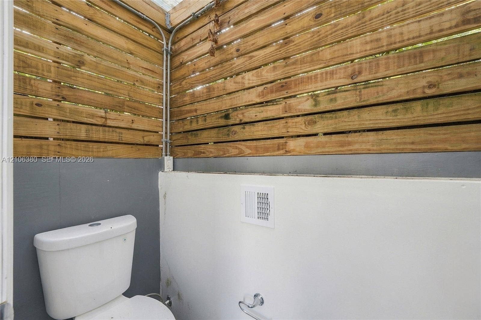 Privacy-fencing enclosure with horizontal wood slats, exposed conduit, and a white porcelain toilet