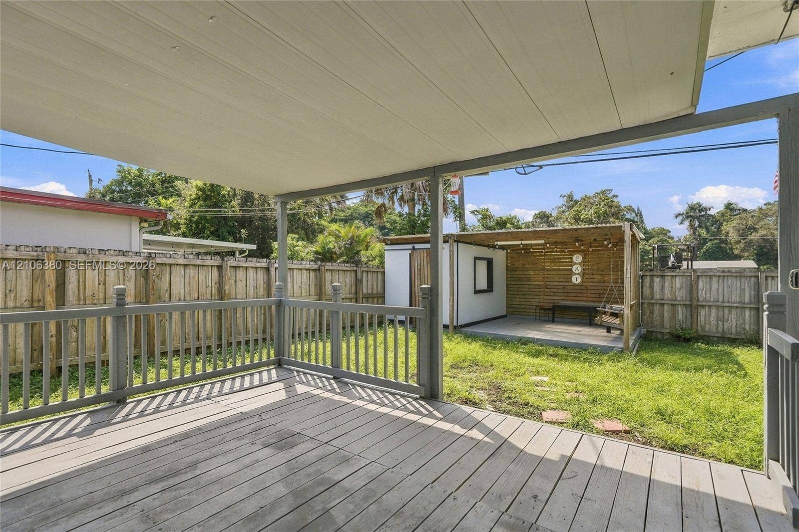 Covered wooden deck featuring a perimeter railing, overlooking a fenced backyard with a detached wood-frame structure and a concrete patio
