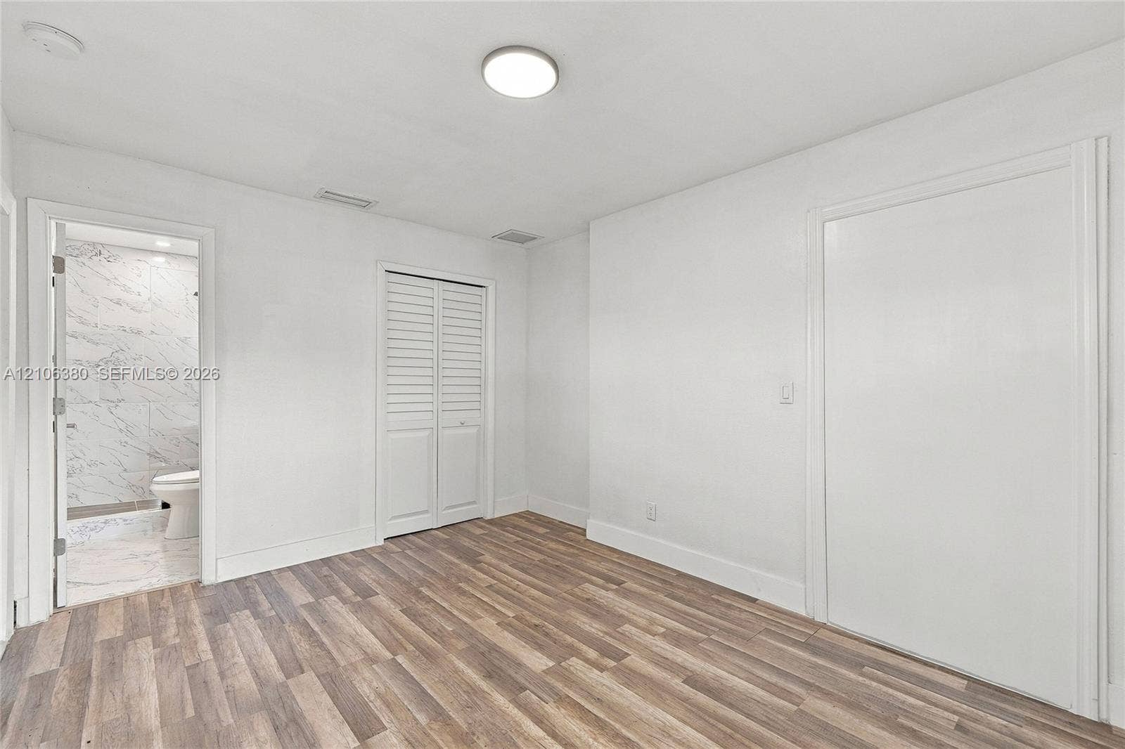 Room featuring wood-finish flooring, white louvered closet doors, a flat-panel door, and a surface-mount ceiling light fixture