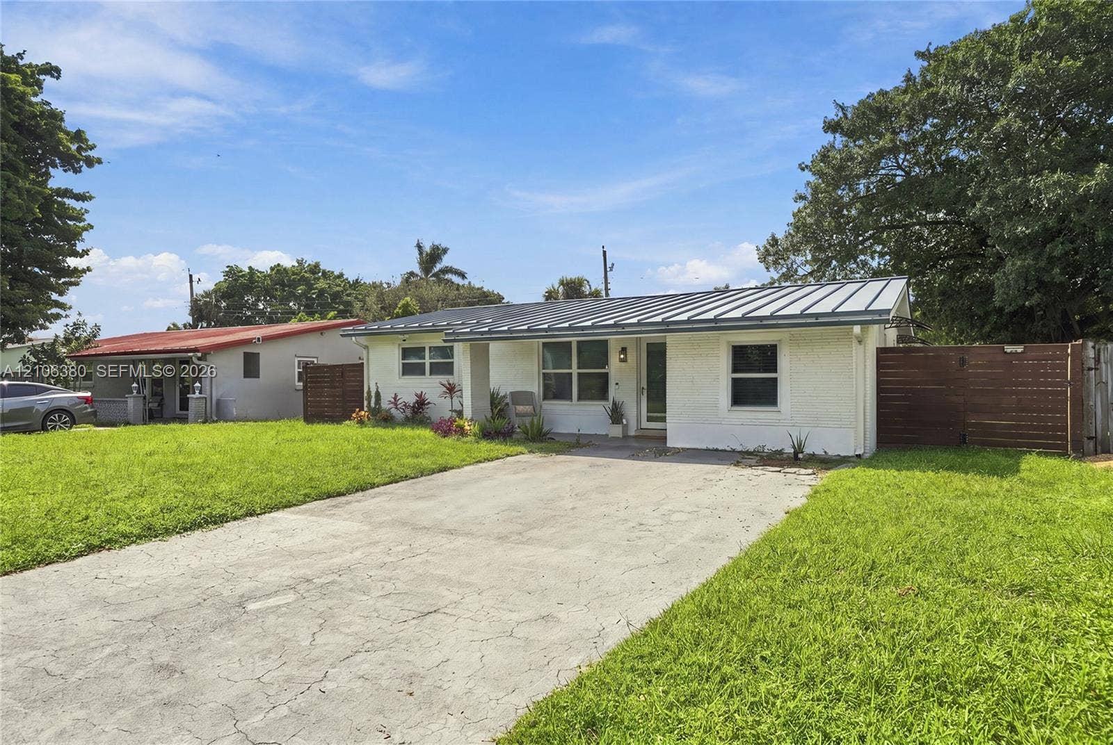 Single-story exterior featuring a painted brick facade, metal roofing, concrete driveway, and wood-slat privacy fencing