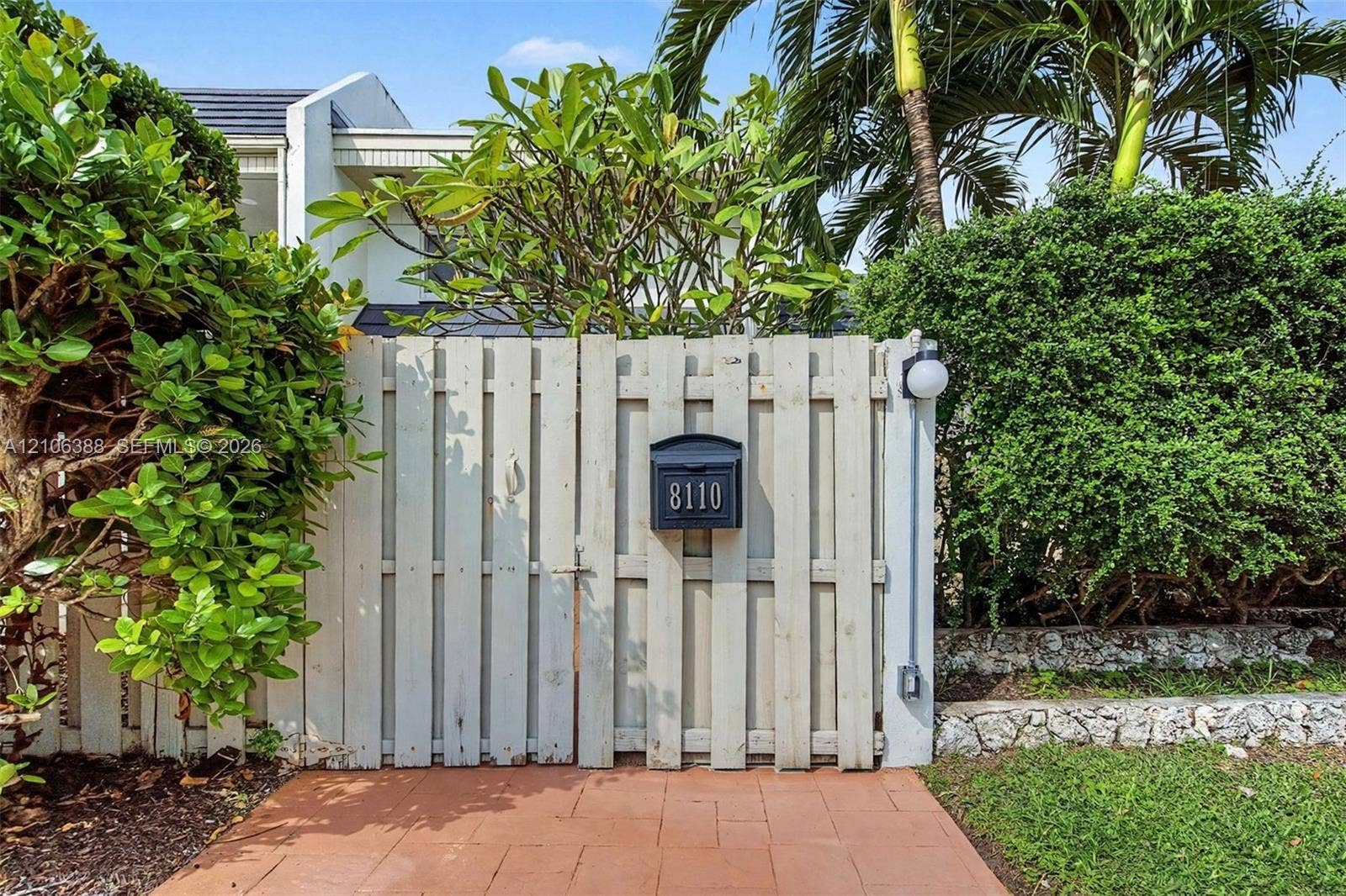 8110 SW 82nd Court, Miami, FL 33143 Vertical wood-plank privacy gate featuring a mounted mailbox and globe-style wall sconce, complemented by terracotta-tile paving and natural stone landscaping borders